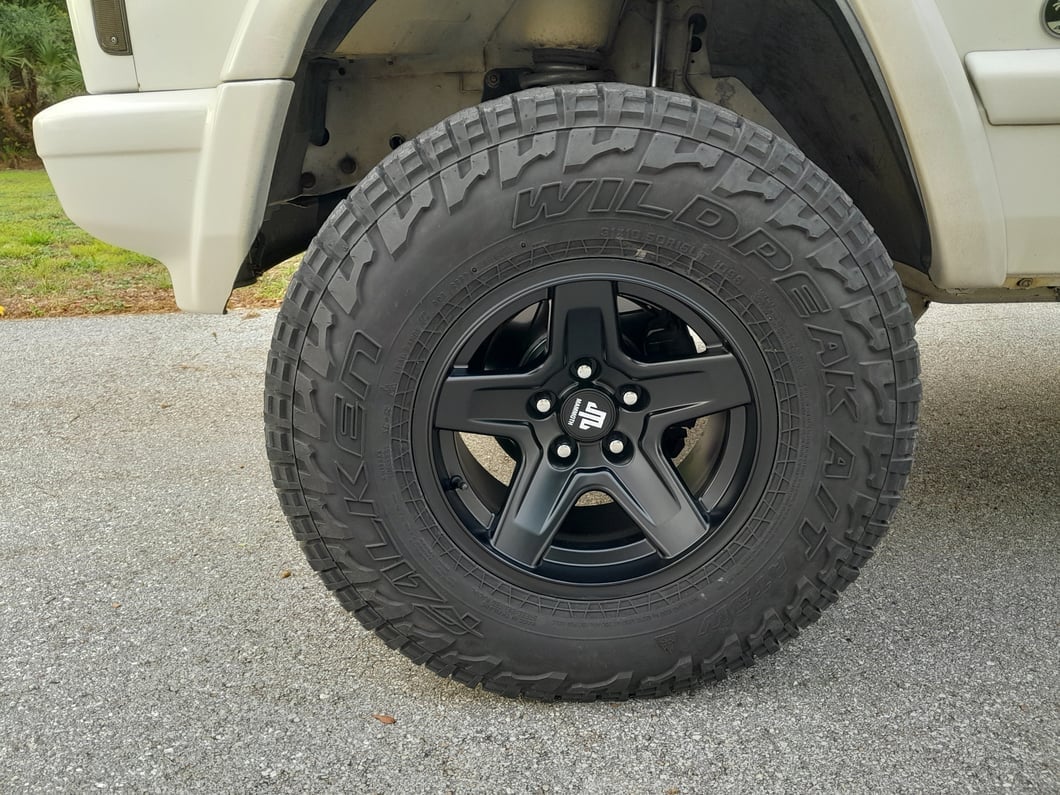 Close-up of a black five-spoke wheel with a Wildpeak A/T tire mounted on a lifted Jeep Cherokee XJ.