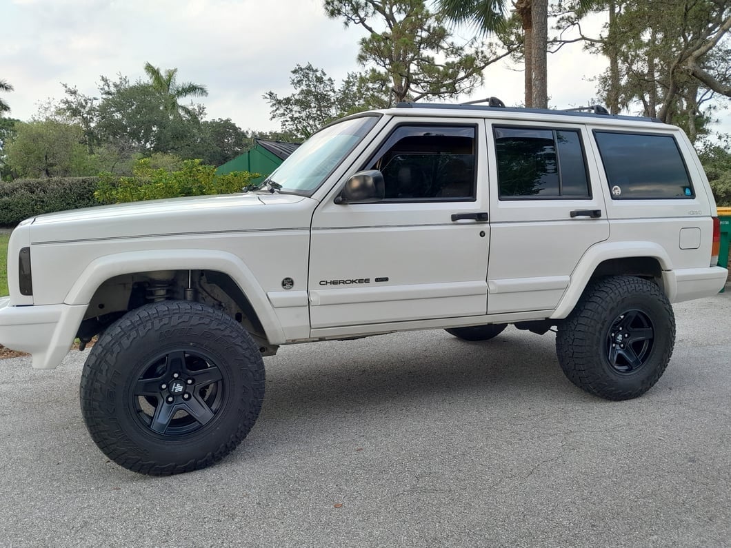 White Jeep Cherokee XJ with a suspension lift, black five-spoke wheels, and all-terrain tires.