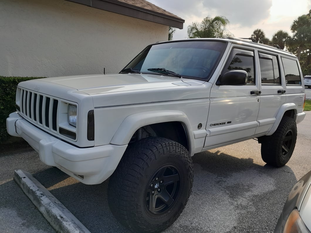 White Jeep Cherokee XJ with a suspension lift, black five-spoke wheels, and all-terrain tires parked in a parking lot.