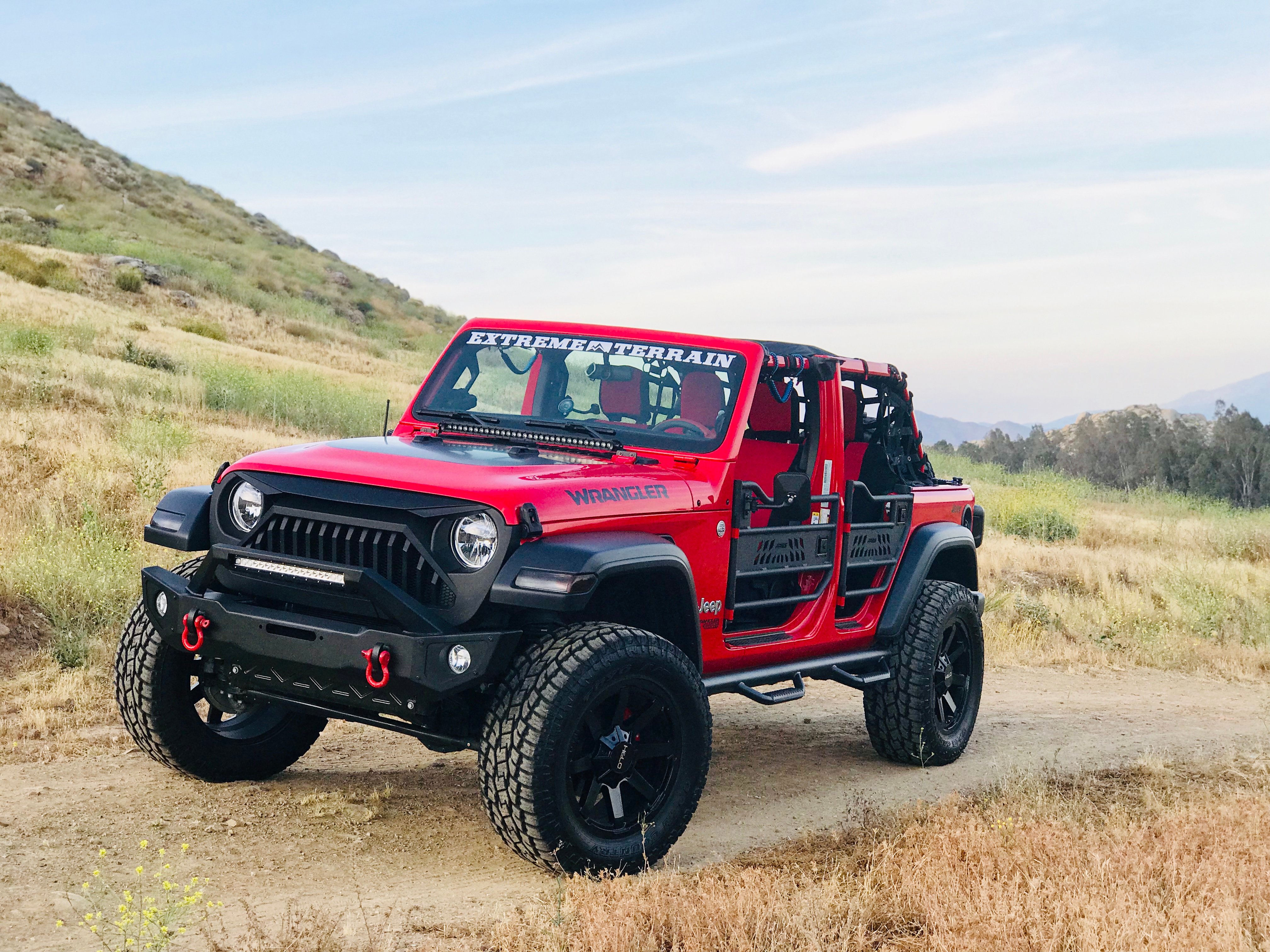 Red Jeep Gladiator with lifted suspension, tube doors, off-road bumper, and all-terrain tires parked on a dirt trail in a grassy hillside landscape.