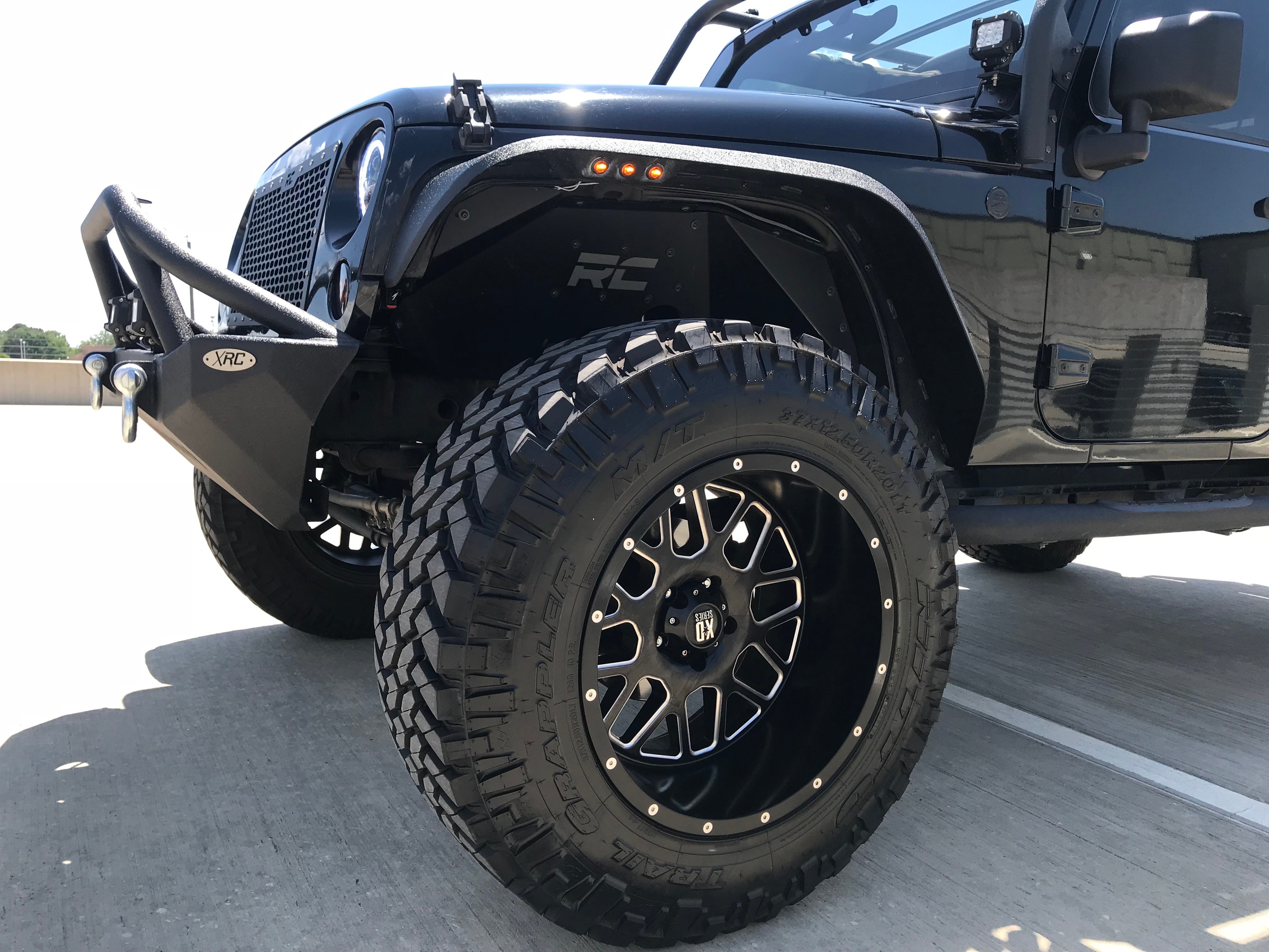 Front three-quarter view of a Jeep Wrangler JK with aftermarket flat fender flares, oversized mud-terrain tires, and an off-road front bumper for increased tire clearance.