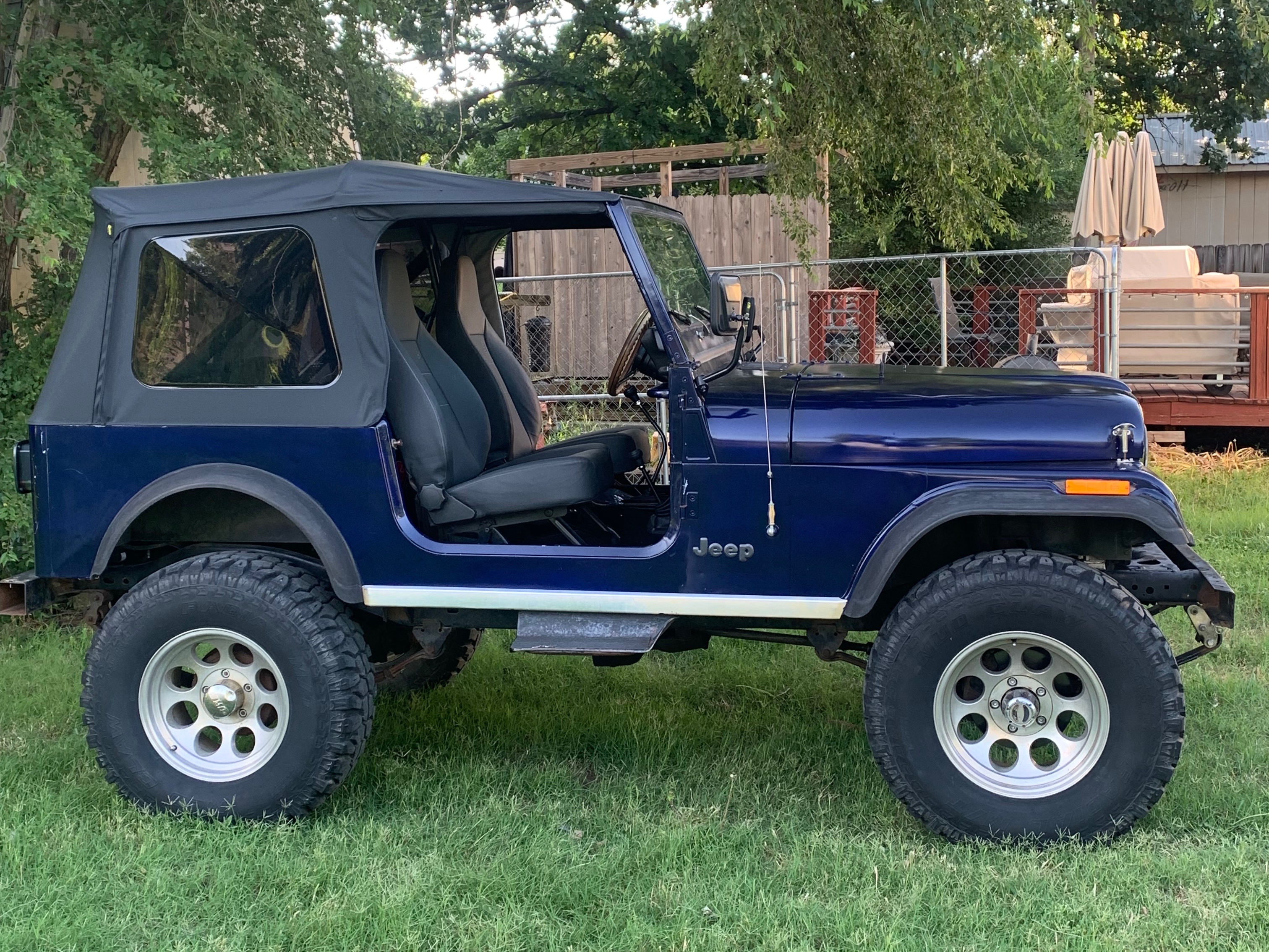 Blue vintage Jeep CJ with oversized off-road tires and soft top parked on grass, featuring an open side cabin and lifted suspension.
