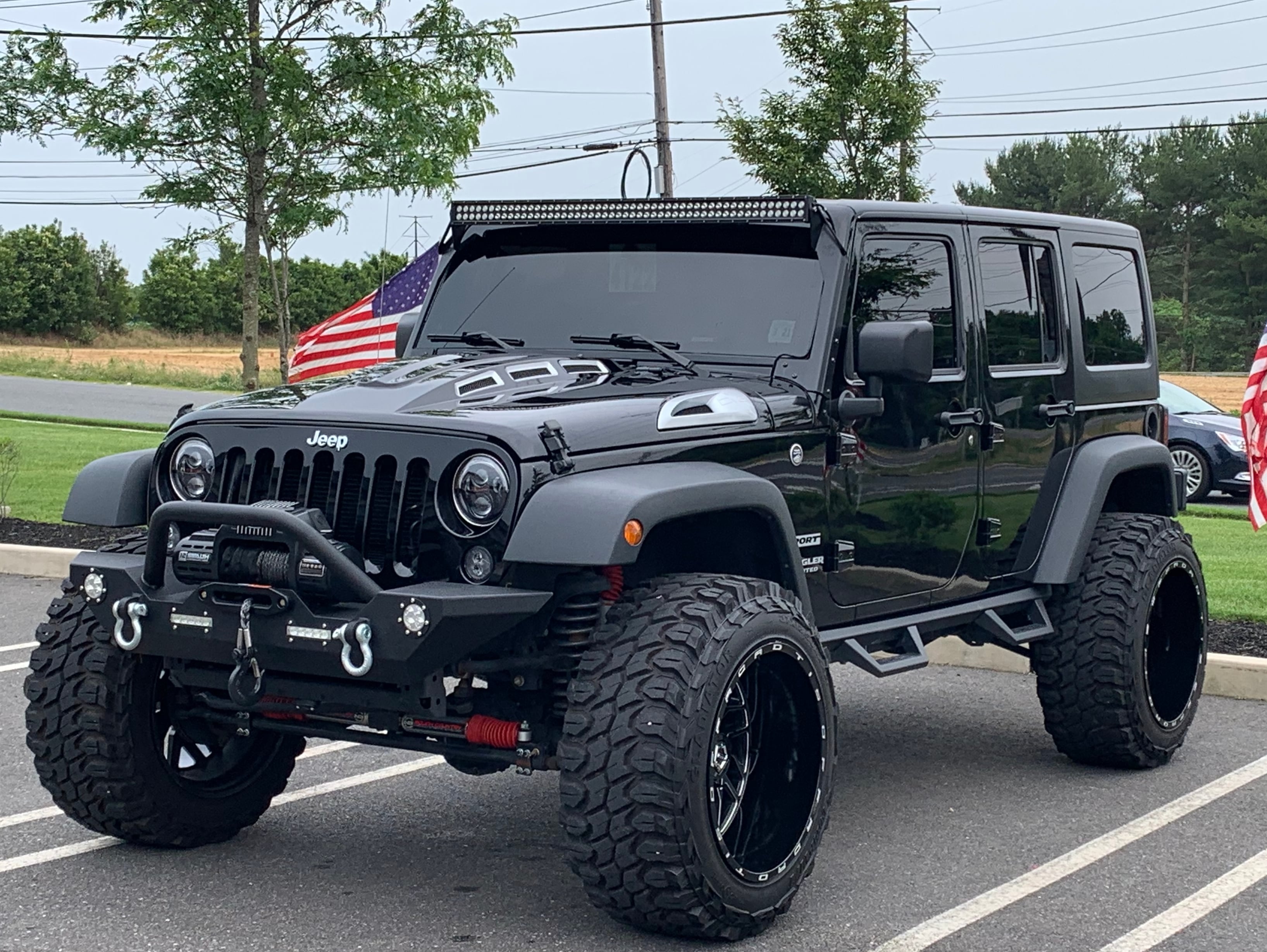 Black lifted four-door Jeep Wrangler with oversized off-road tires, aftermarket bumper, roof light bar, and winch parked in a parking lot.