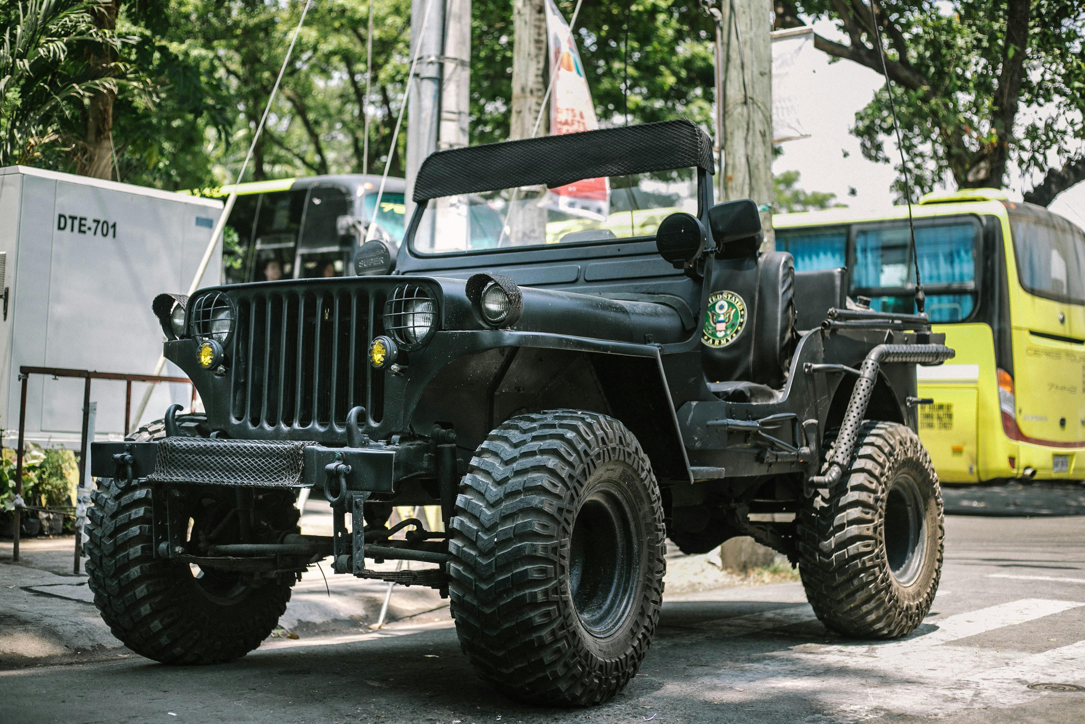 Modified black military-style Jeep with oversized off-road tires parked on a city street, featuring an open cabin, rugged front grille, and lifted suspension.