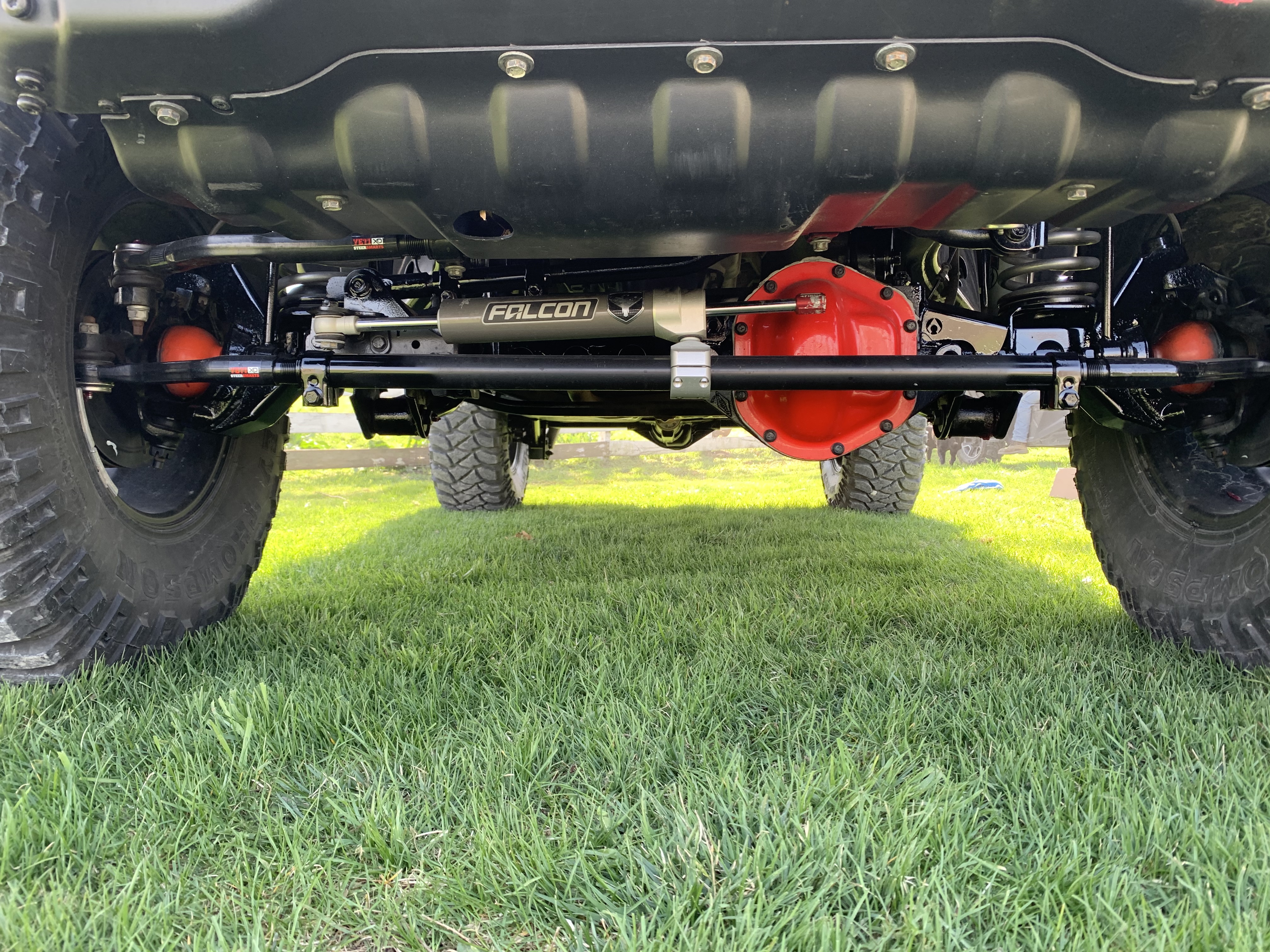 Undercarriage view of a lifted off-road vehicle showing upgraded suspension components, heavy-duty axle housing, steering stabilizer, and oversized all-terrain tires.