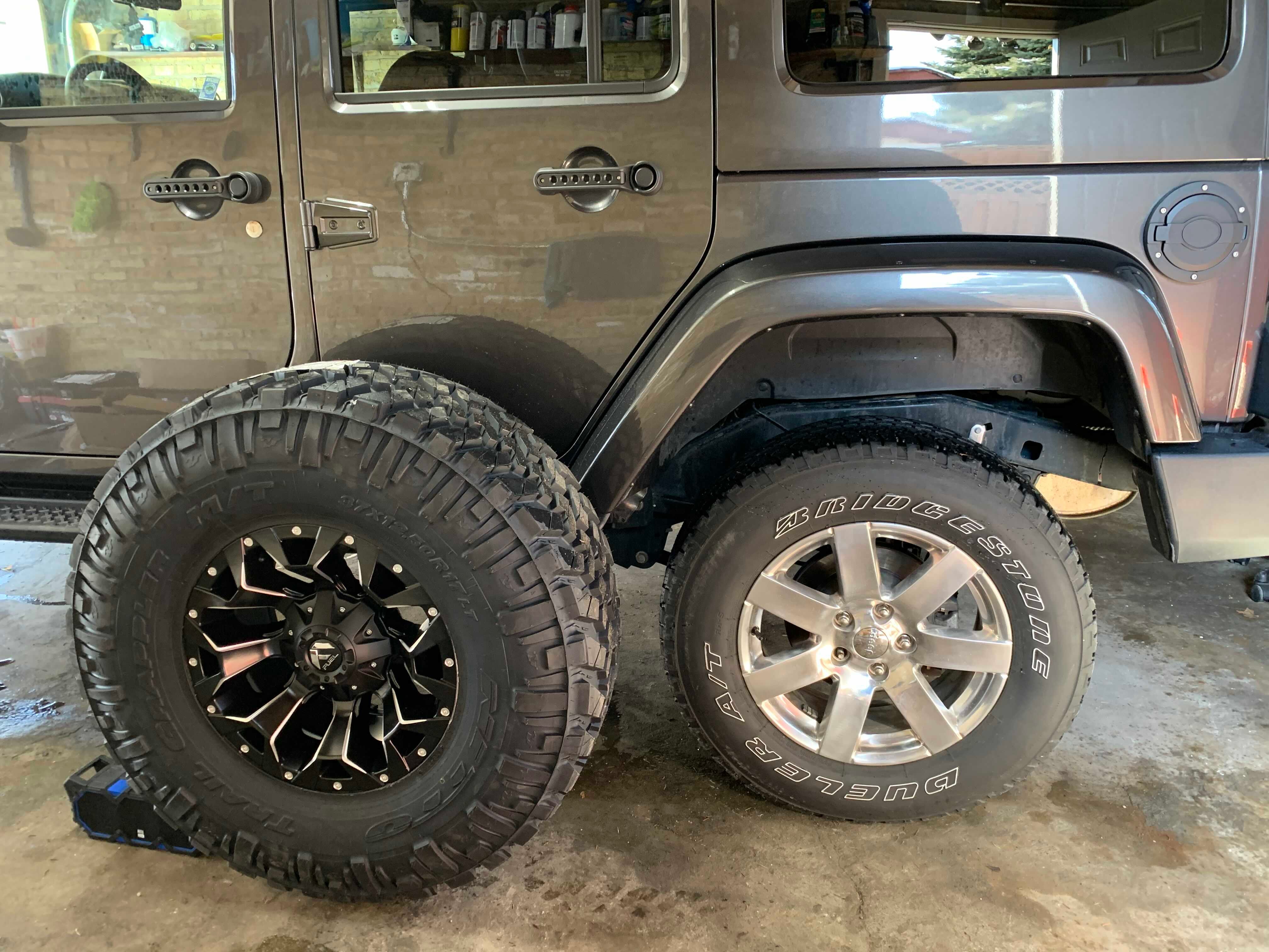 Gray Jeep Wrangler with oversized off-road tire and aftermarket wheel displayed beside the factory tire for size comparison in a garage.