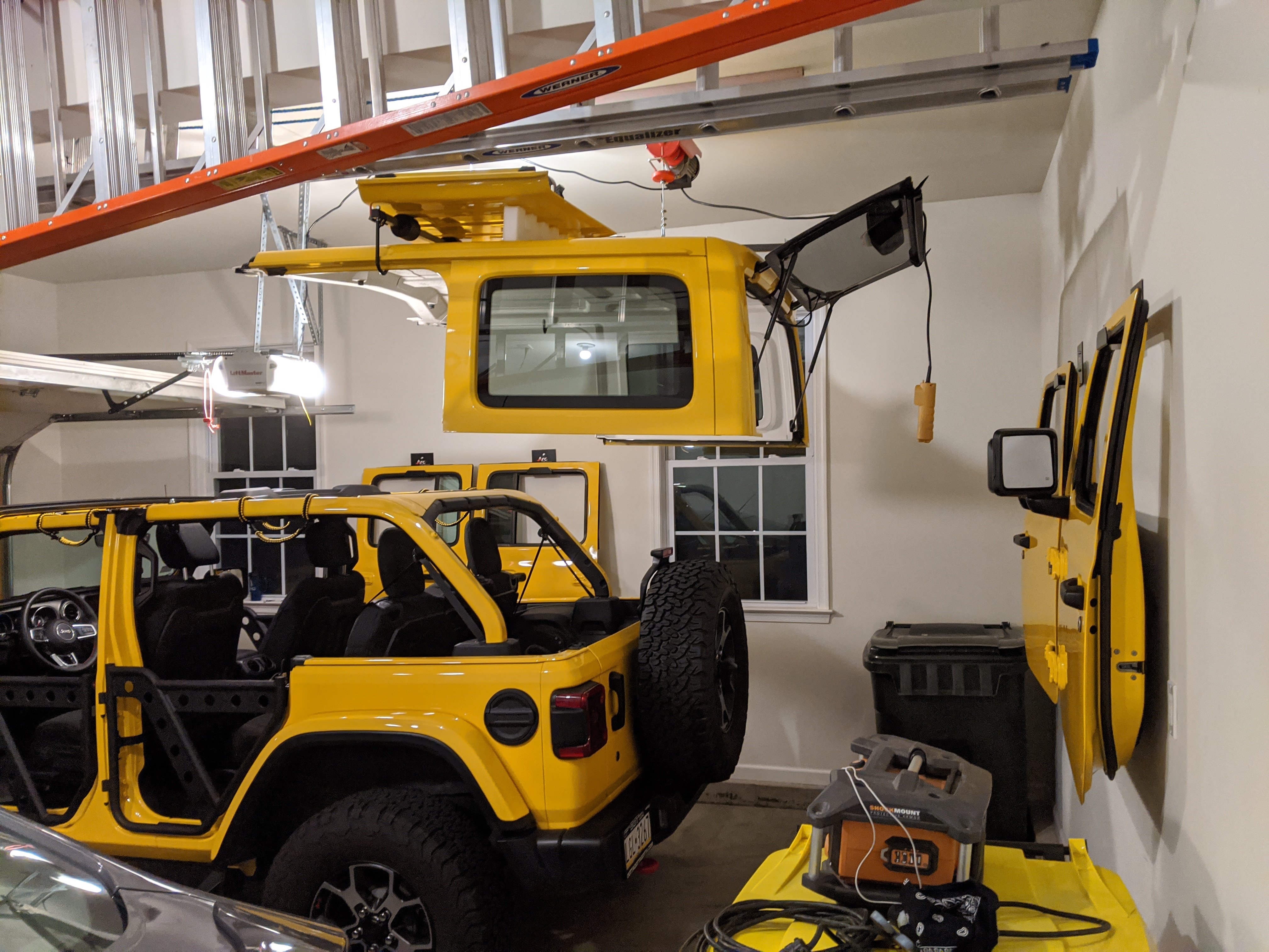 Yellow Jeep Wrangler with the hardtop suspended from a ceiling-mounted garage hoist, showing the removable doors stored on the wall and the open-air vehicle ready for driving.